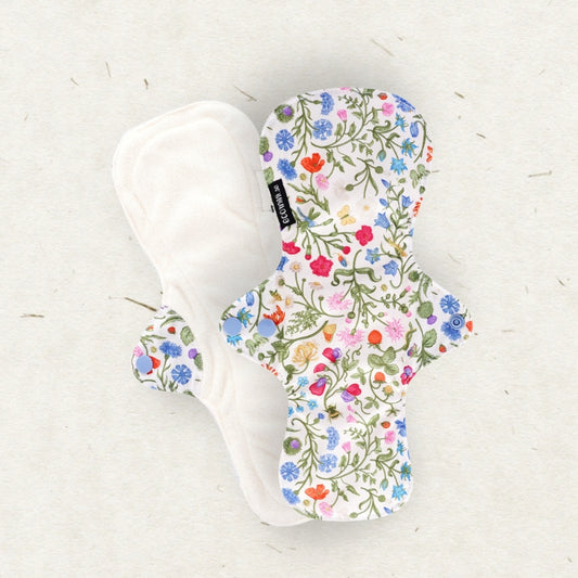 Two reusable cloth menstrual pads on a light textured background. One pad is shown from the back in solid white fabric, and the other is shown from the front with a dense multicolor floral print featuring green leaves and red, blue, pink, and yellow flowers on a white base.