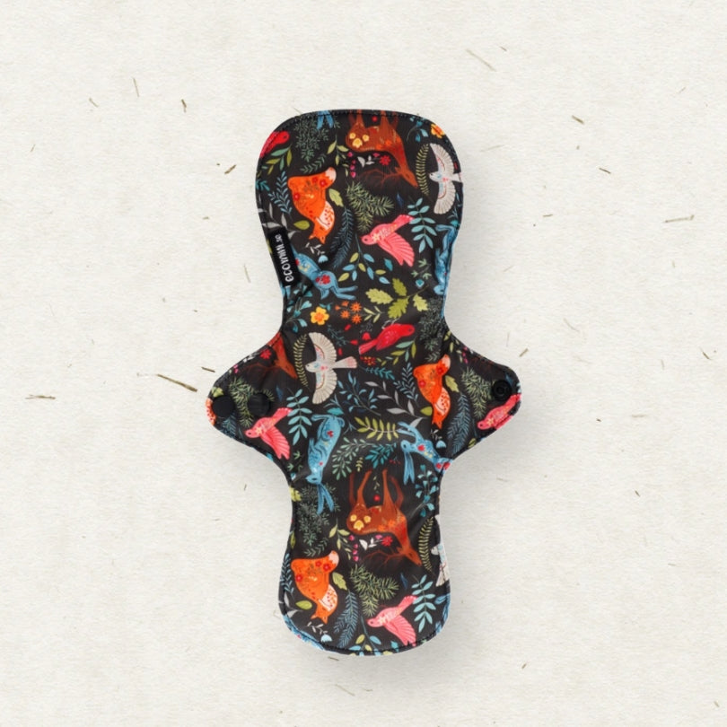 A reusable menstrual pad with a dark background and a colorful pattern of birds, foxes, and leaves, shown on a textured off-white surface.