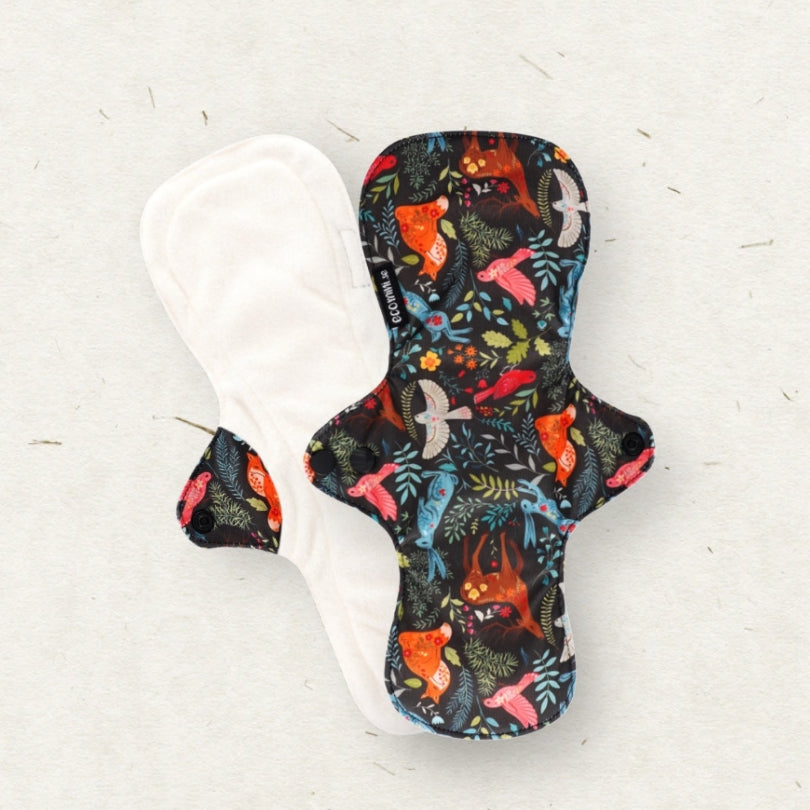 Two reusable cloth menstrual pads on a light textured background. One pad is shown from the patterned side, featuring a dark background with a colorful print of foxes, birds, and leaves.