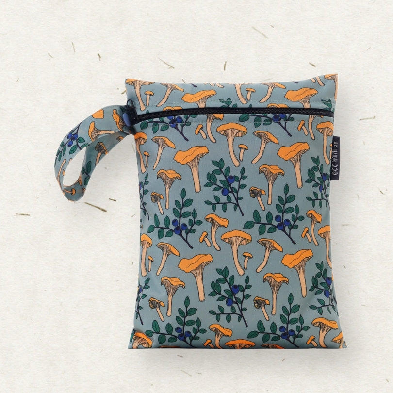 Eco Mini brand wet bag with a matching wrist strap, featuring an all-over print of orange chanterelle mushrooms and green leaves on a muted blue-gray background. The bag is laid flat on a textured off-white surface.