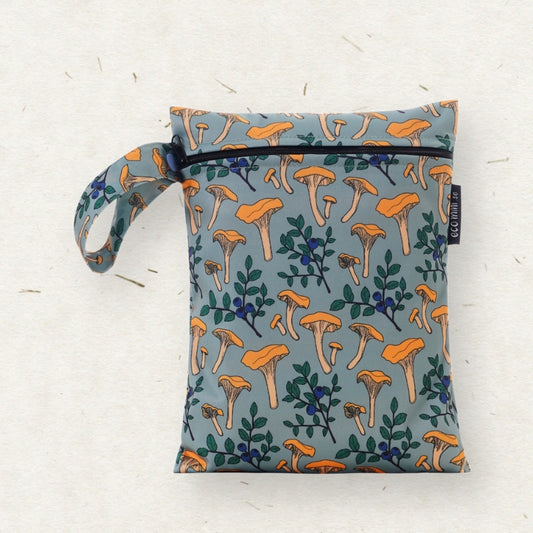 Eco Mini brand wet bag with a matching wrist strap, featuring an all-over print of orange chanterelle mushrooms and green leaves on a muted blue-gray background. The bag is laid flat on a textured off-white surface.