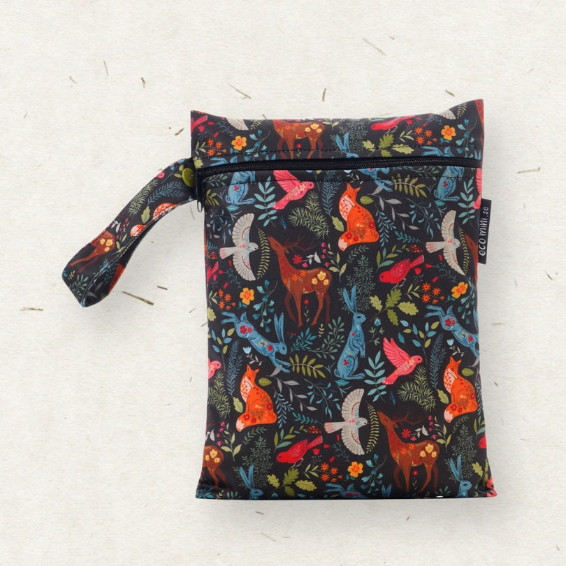 A reusable wet bag with a handle, featuring a colorful forest animal print on a black background. The pattern includes orange foxes, blue birds, white birds, and green foliage.