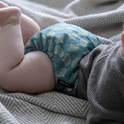 A close-up of a baby's legs and torso lying on a textured gray blanket, wearing a blue reusable cloth diaper with a beige ginkgo leaf pattern. A small black label on the diaper reads 'ECO MINI'.