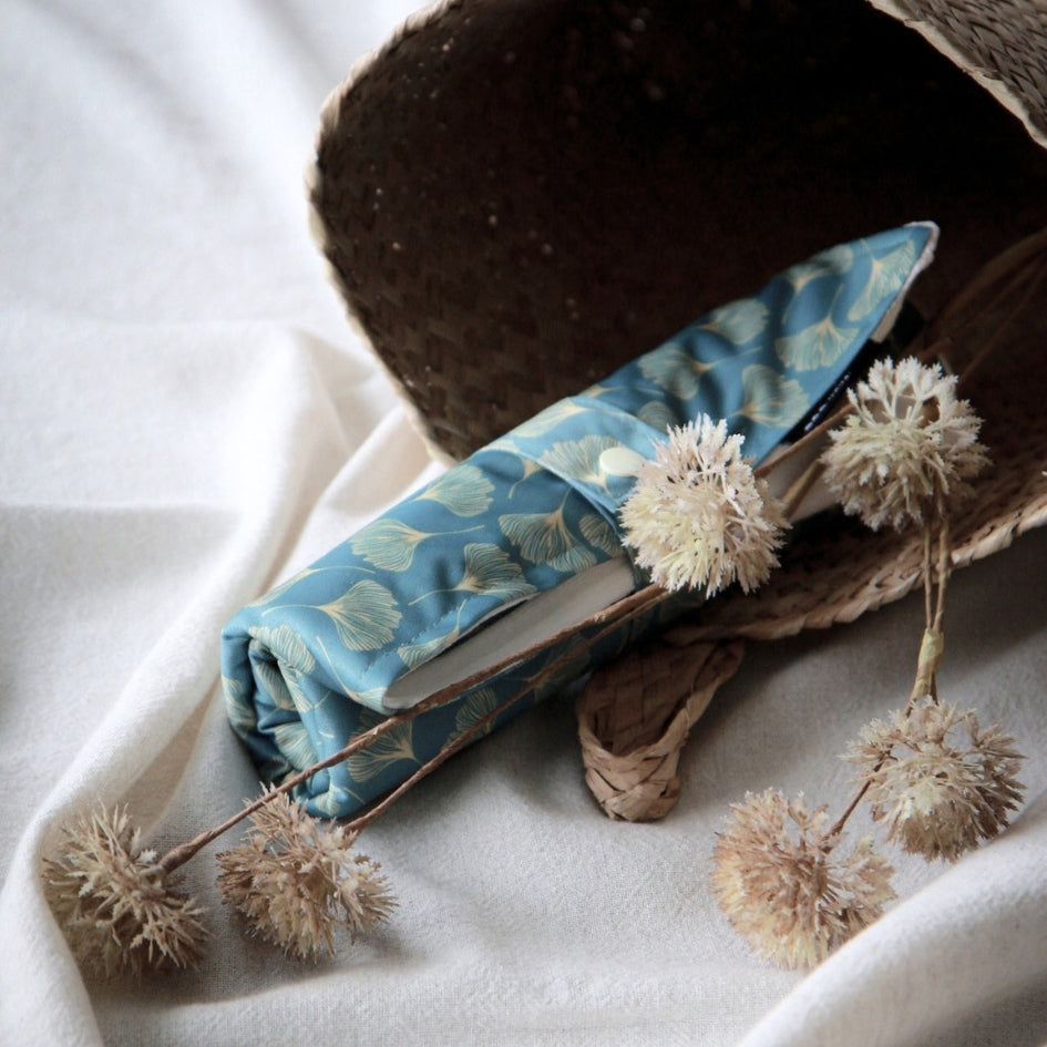 A rolled change mat in light blue with a pale botanical leaf pattern, placed on white draped fabric beside dried bunny tail flowers and a woven hat.