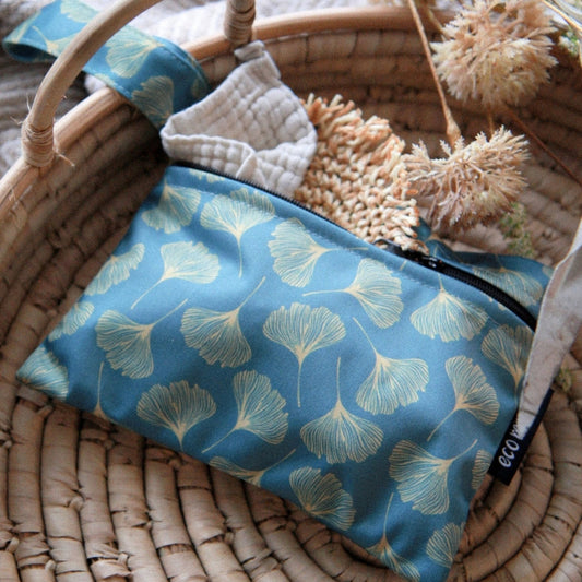 A blue fabric wet bag with a light beige ginkgo leaf pattern, resting inside a woven wicker basket with dried flowers and a small white textured item.