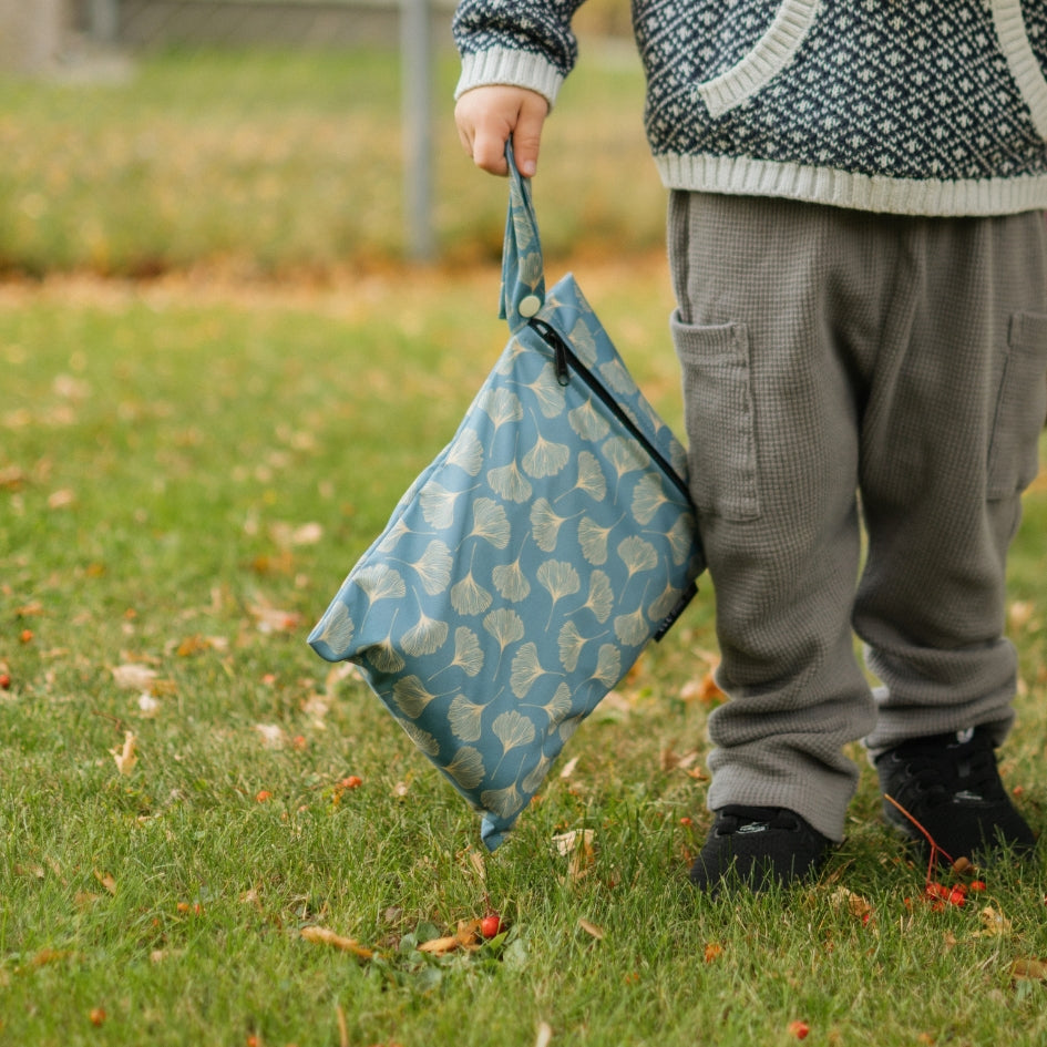 A child standing on grass outdoors holds up a reusable blue fabric wet bag with a white leaf pattern. The child is wearing gray waffle-knit pants and a patterned knit sweater.