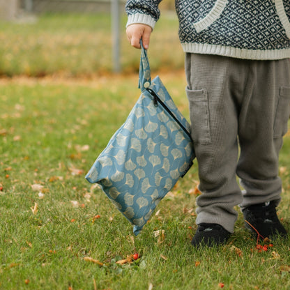 A child standing on grass outdoors holds up a reusable blue fabric wet bag with a white leaf pattern. The child is wearing gray waffle-knit pants and a patterned knit sweater.
