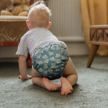 A baby crawling on a carpeted floor, viewed from behind, wearing a white t-shirt and a blue patterned cloth diaper.