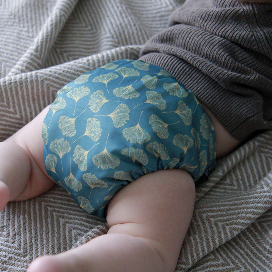 A close-up of a baby wearing a teal blue reusable cloth diaper with a light green ginkgo leaf pattern, lying on a textured gray and white herringbone blanket.
