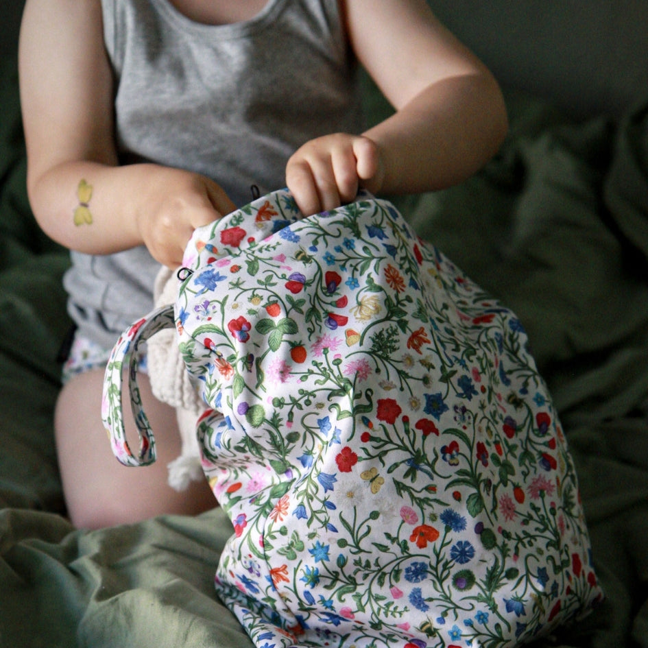 A child in a gray tank top holds a fabric wet bag with a colorful floral print on a green bed.