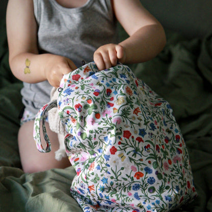 A child in a gray tank top holds a fabric wet bag with a colorful floral print on a green bed.