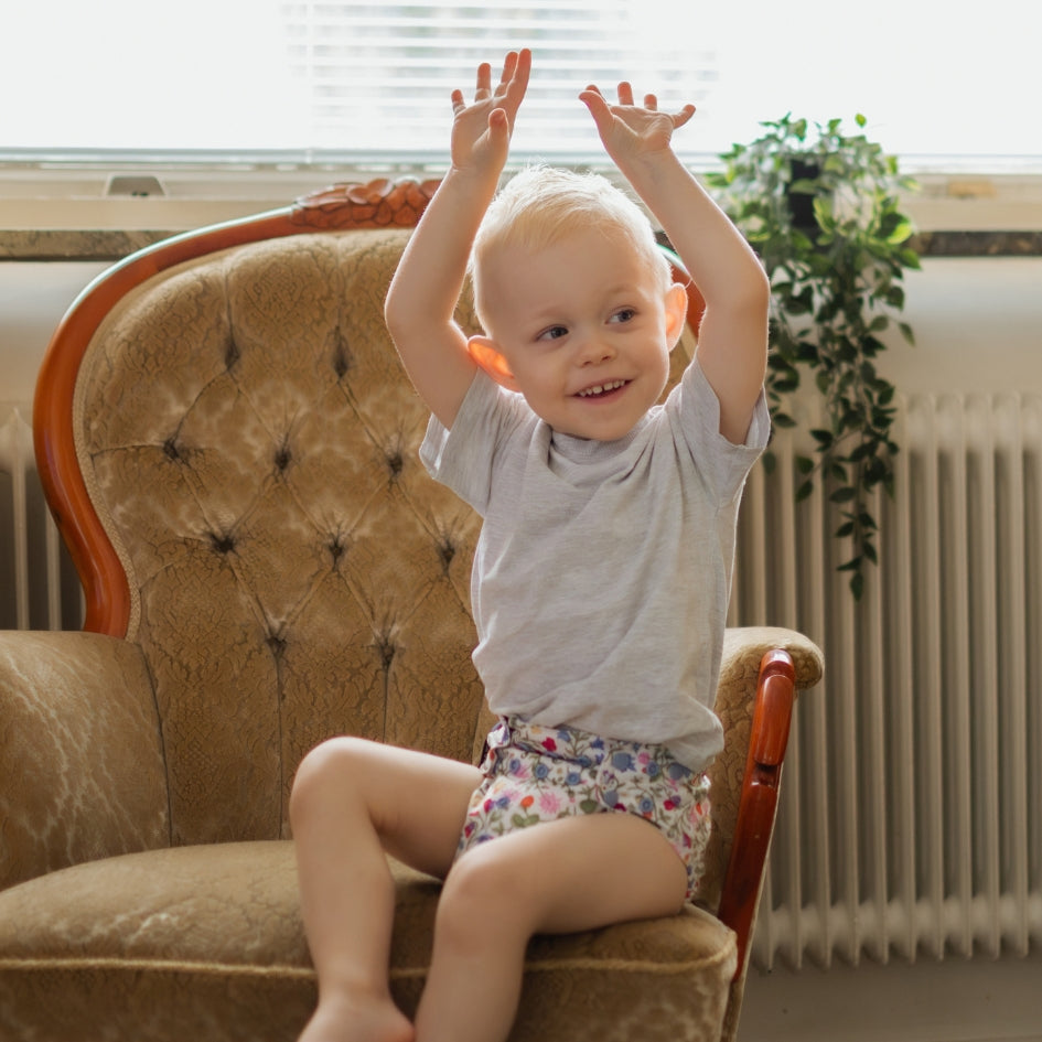 A smiling toddler with blonde hair sitting on a tufted brown armchair, wearing a light gray t-shirt and floral patterned diaper cover, with their hands raised in the air.