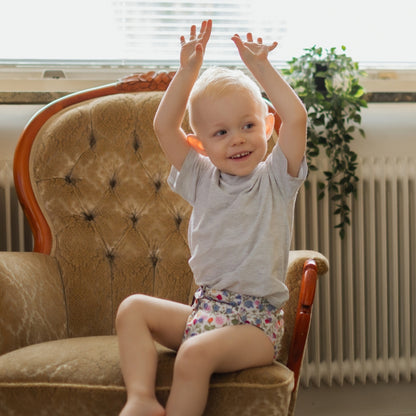 A smiling toddler with blonde hair sitting on a tufted brown armchair, wearing a light gray t-shirt and floral patterned diaper cover, with their hands raised in the air.