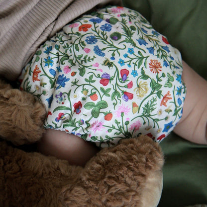 A close-up of a baby's legs and torso wrapped in a white cloth diaper covered in a colorful floral and botanical print, resting on a brown teddy bear and green fabric.