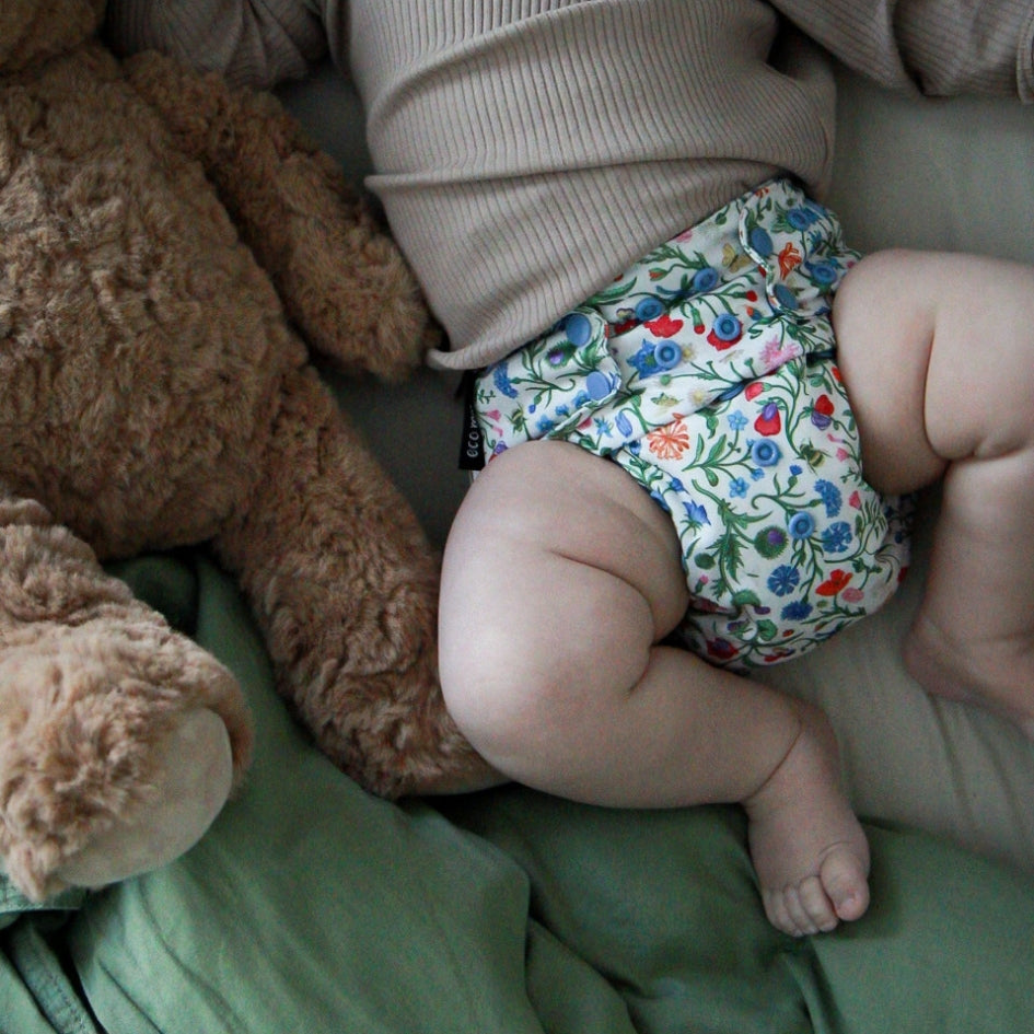 A close-up lifestyle shot of a baby wearing a white reusable cloth diaper with a colorful floral print, lying next to a brown teddy bear on a green blanket.