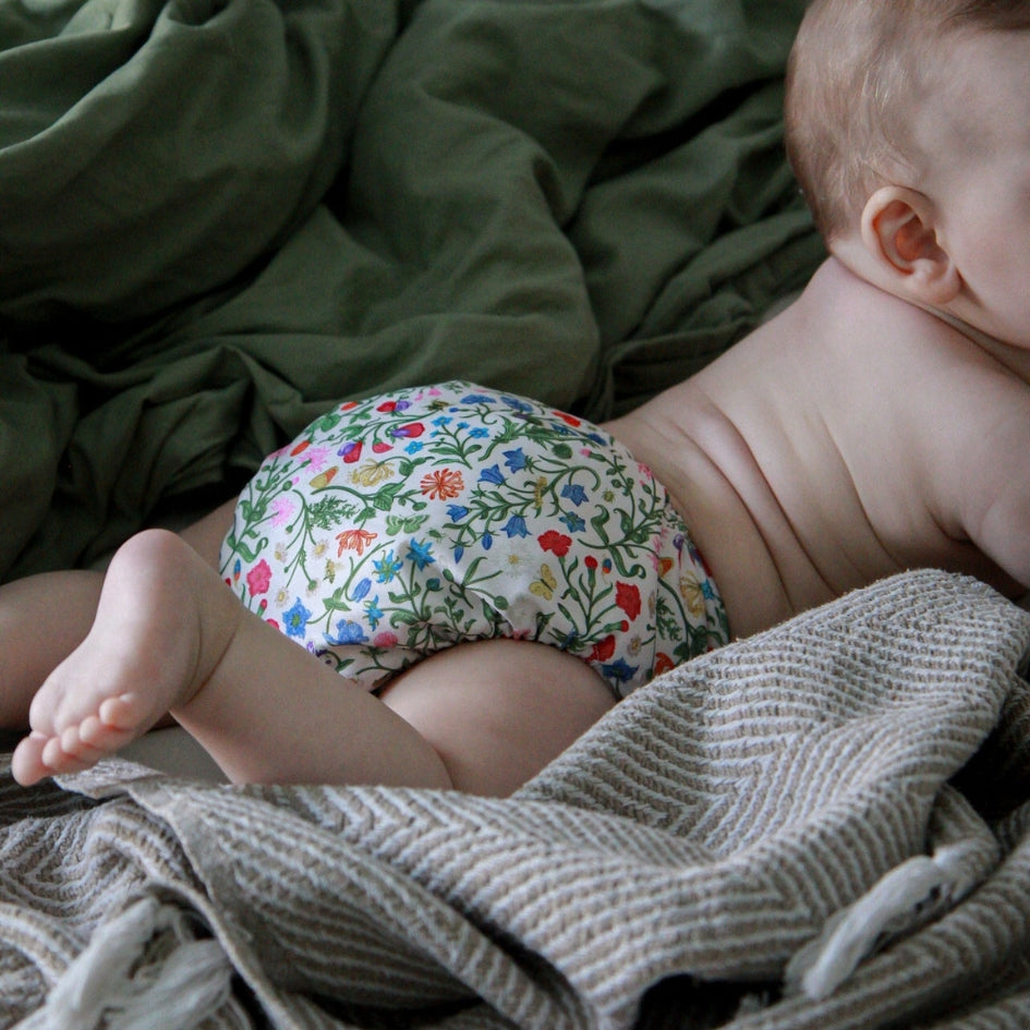 A baby lying on a bed wearing a floral print reusable cloth diaper, with green and gray blankets in the background.