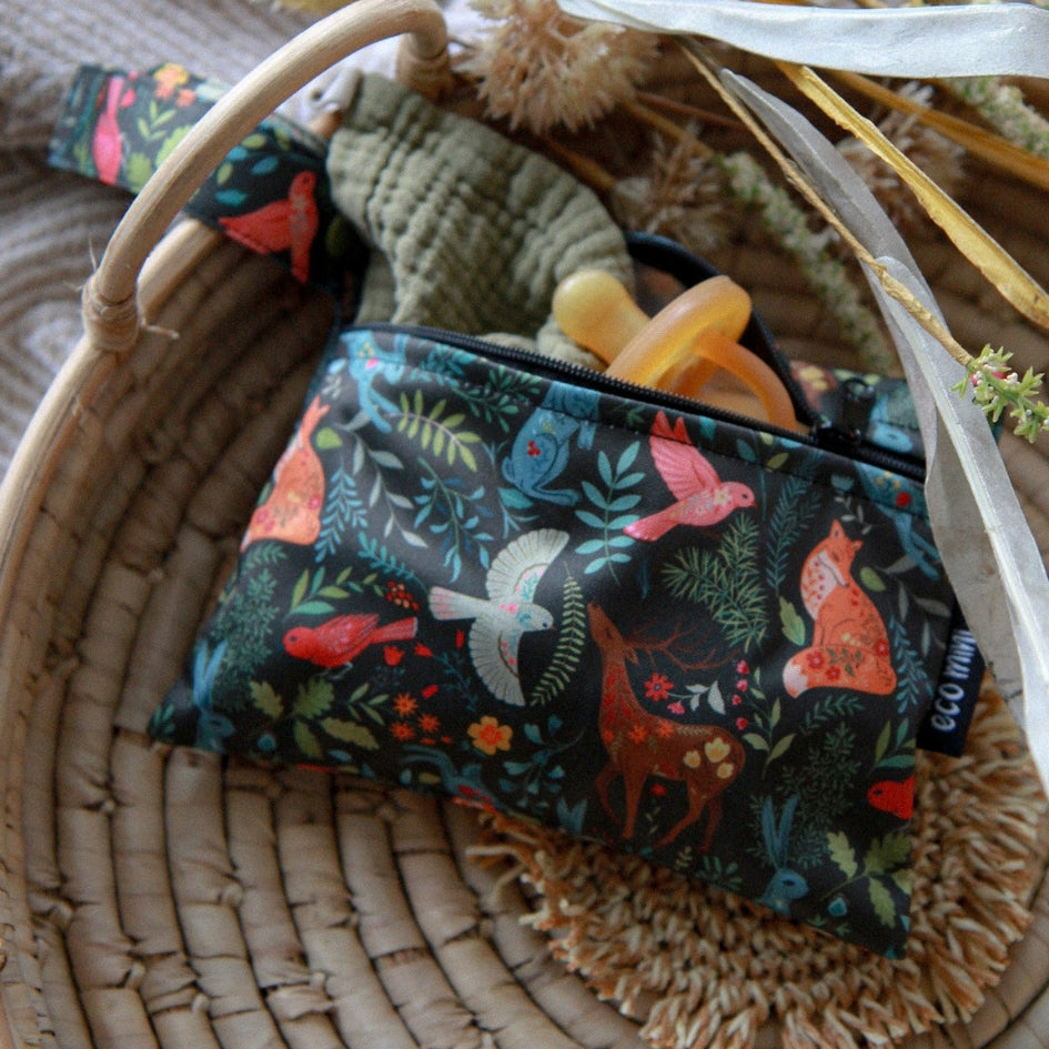 A wet bag with a colorful forest animal print, shown inside a woven basket with baby accessories like a muslin cloth and pacifier.
