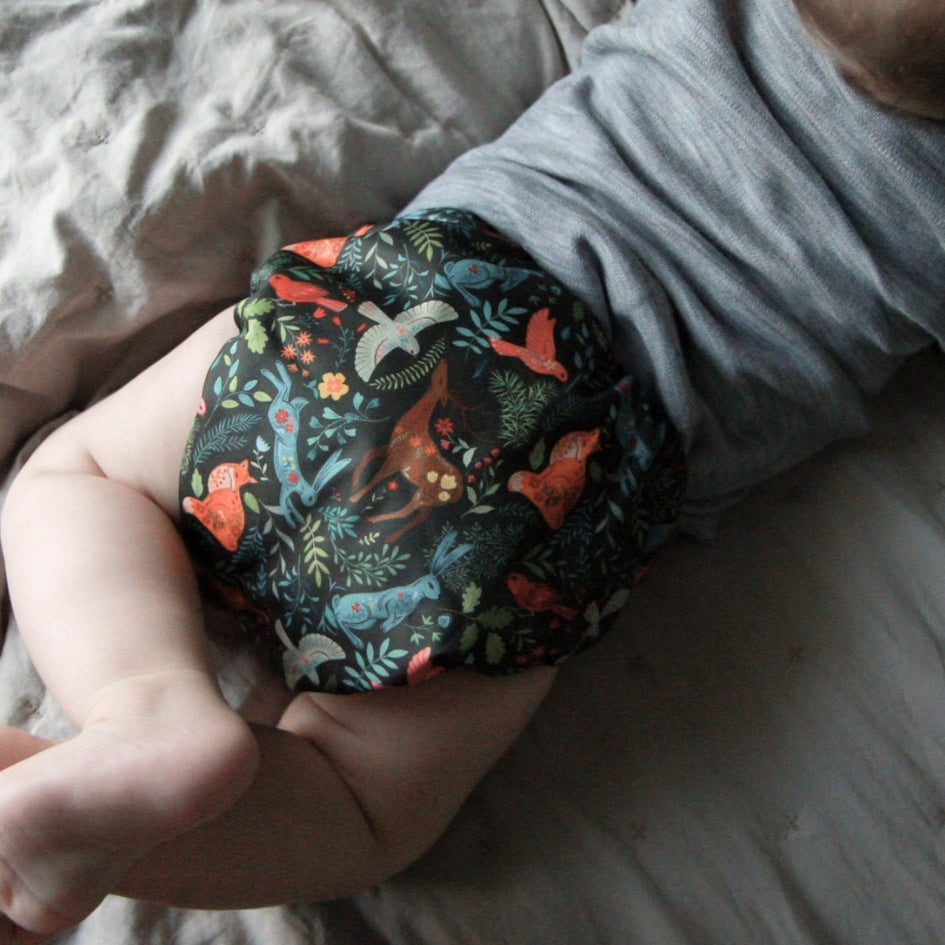 A baby wearing a dark patterned cloth diaper with colorful woodland animals and foliage, paired with a gray shirt, lying on a bed.