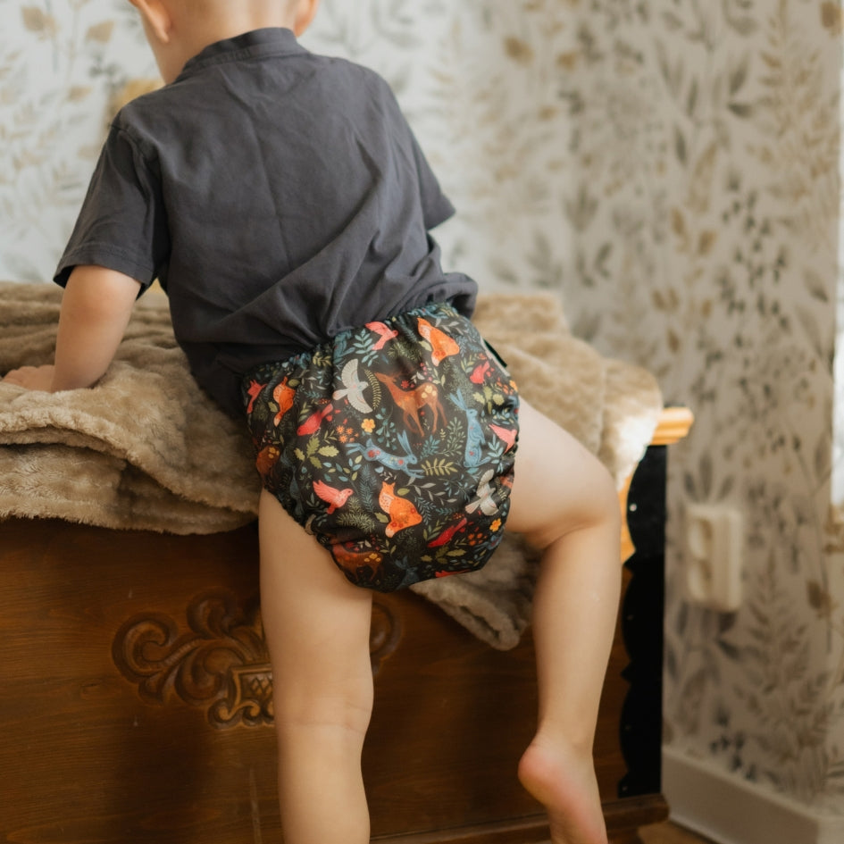 A child seen from behind wearing a dark gray t-shirt and a highly colorful, patterned reusable cloth diaper while sitting on a wooden bed in a home setting.