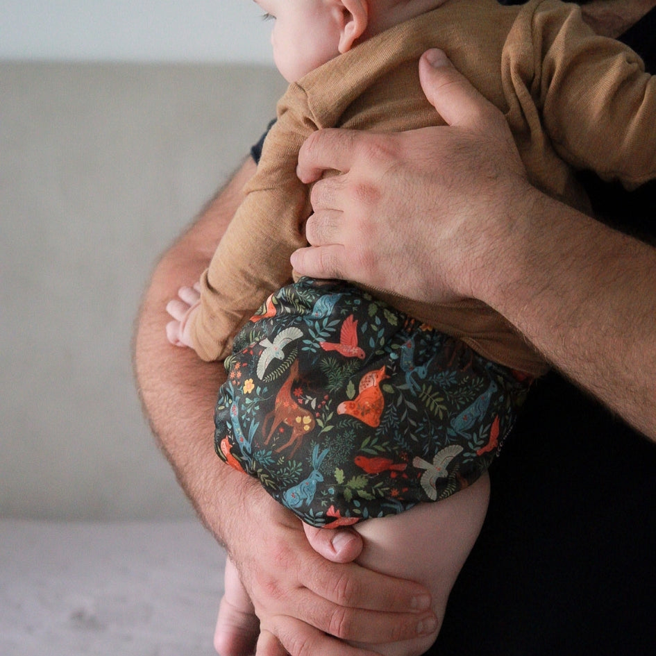 A baby wearing a dark patterned cloth diaper with colorful birds and foliage, held by an adult's arms. The baby is also wearing a tan long-sleeve top.